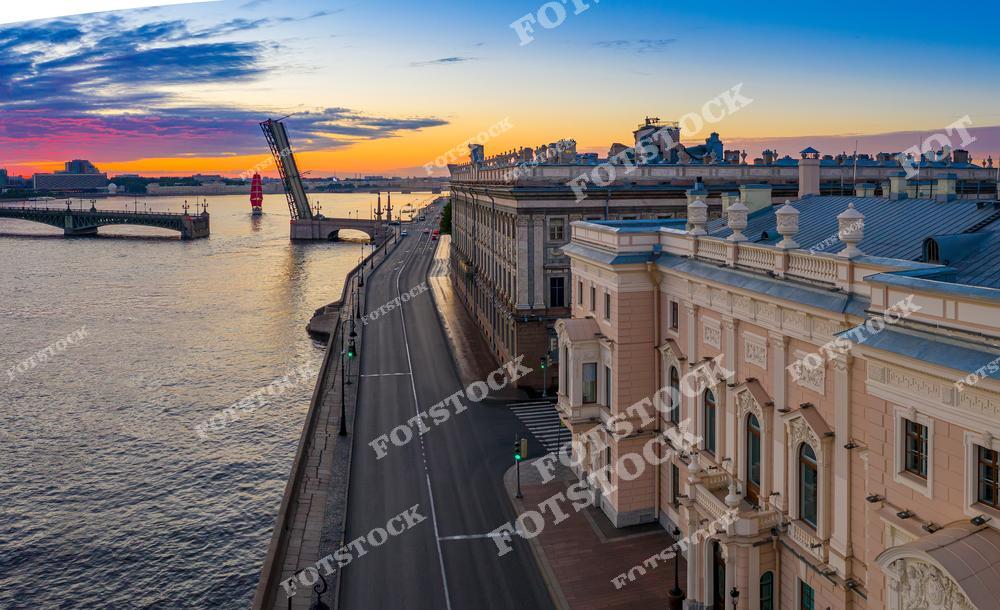 На фотографии показан центр Санкт-Петербурга в период белых ночей. Снимок сделан сверху и захватывает пустынную набережную Невы, разведенный Троицкий мост, корабль с алыми парусами и красивое небо.